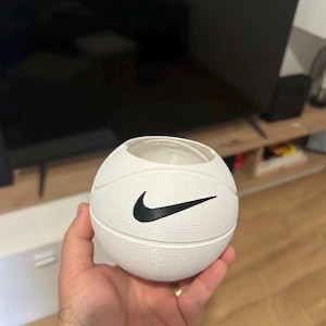 Pode incluir: Um recipiente branco em forma de bola de basquete com um logotipo Nike preto. O recipiente tem uma superfície texturizada e um topo oco. O recipiente é segurado na mão de uma pessoa.