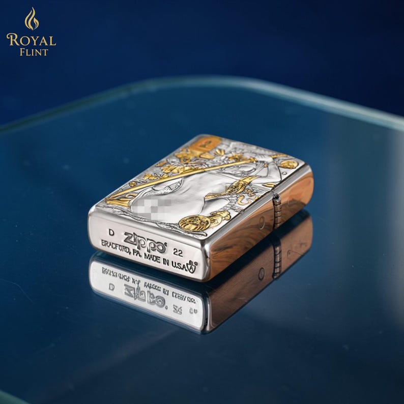 May include: A silver and gold Zippo lighter with an engraved design. The lighter has a rectangular shape with rounded edges. The text "Zippo 22" and "Bradford, PA. Made in USA" is visible on the bottom. The lighter is reflected on a blue surface.