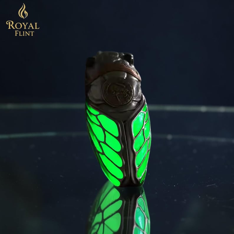 May include: A unique cicada-shaped lighter with glowing green wings. The lighter has a dark metallic body with a brown head and intricate detailing. The text "ROYAL FLINT" and a flame logo are visible at the top.