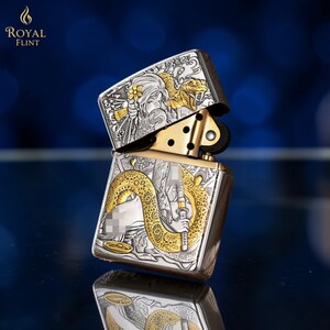 May include: Silver and gold-toned metal lighter with intricate engraved designs. The lighter features a detailed illustration of a woman, a snake, and floral accents. The top of the lighter is open, revealing the internal mechanism. The brand name "ROYAL FLINT" is visible.