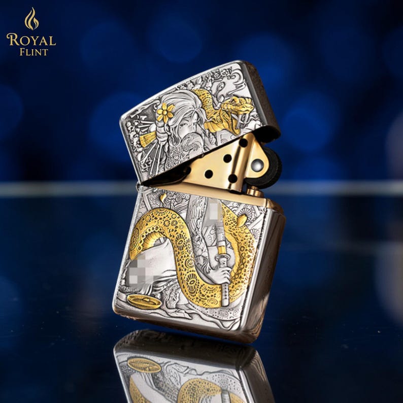 May include: Silver and gold-toned metal lighter with intricate engraved designs. The lighter features a detailed illustration of a woman, a snake, and floral accents. The top of the lighter is open, revealing the internal mechanism. The brand name "ROYAL FLINT" is visible.