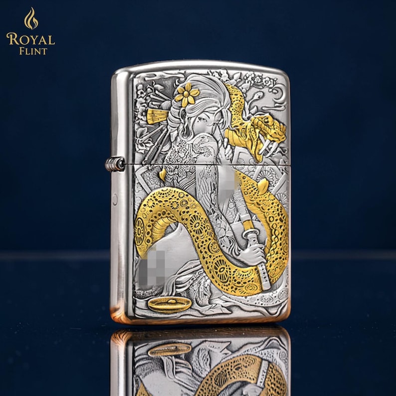 May include: Silver-toned metal lighter with a detailed engraved design. The design features a woman with a flower in her hair, a golden snake, and a sword. The lighter has a hinged lid and a classic rectangular shape.
