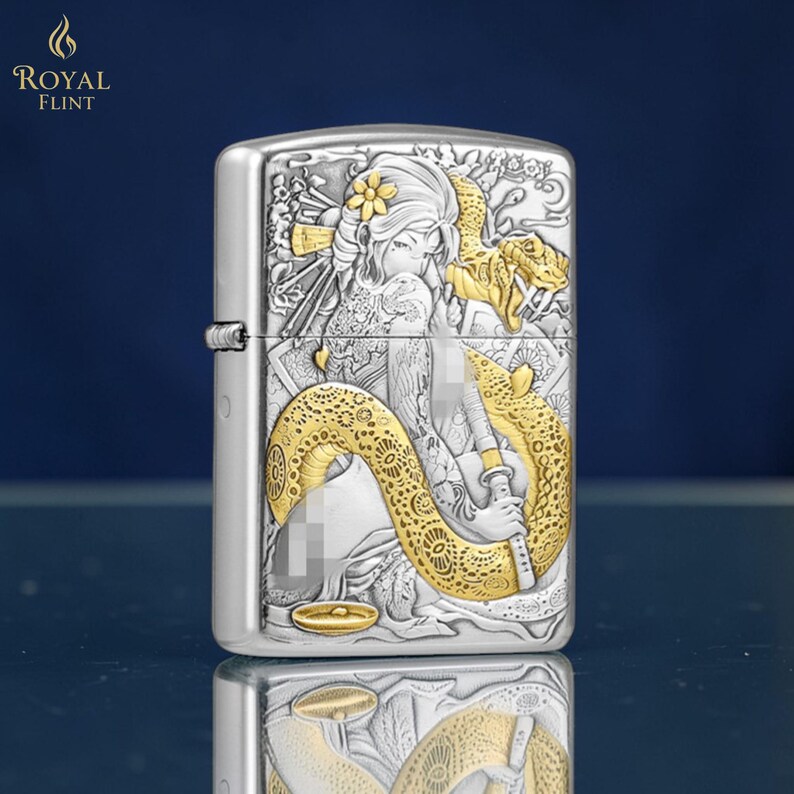 May include: Silver-toned lighter with an engraved design featuring a woman and a golden snake. The lighter has a rectangular shape and a hinged lid. The design includes floral accents and the words "ROYAL FLINT".