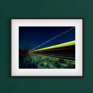 Peut inclure: Tirage encadré présentant une photographie à longue exposition d'un train de nuit. L'image capture des traînées de lumière jaunes, rouges et blanches sur un ciel bleu foncé, avec des voies ferrées et un sol enneigé au premier plan.