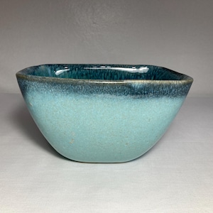 May include: A square ceramic bowl with a turquoise and dark blue glaze. The bowl has a smooth, glossy finish and a slightly textured surface. The interior is a darker shade of blue, and the exterior is a lighter turquoise.