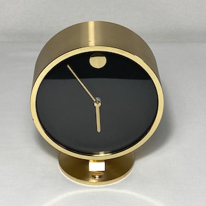 May include: A gold-toned, cylindrical clock with a black face and gold hands. The clock has a circular base and a small, round detail at the top. The clock's design is minimalist and modern.