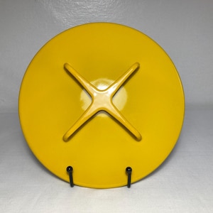 May include: A bright yellow, round plate with a unique starburst design on the back. The plate is held up by two black metal stands. The plate's design features four curved arms extending from the center.