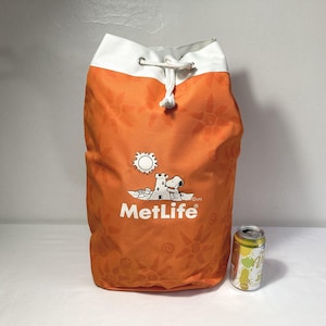 May include: Orange drawstring bag with a white top and a MetLife logo featuring Snoopy. The bag has a repeating sun and flower pattern. A can of soda is next to the bag.