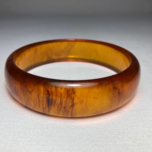 May include: A vintage, amber-colored bangle bracelet. The round bracelet has a marbled appearance with dark brown streaks throughout the translucent material. The surface is smooth and glossy, reflecting light.