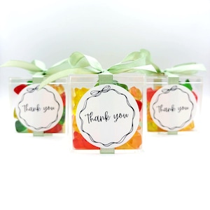 May include: Three clear square plastic boxes filled with colorful gummy candies. Each box is tied with a light green ribbon and features a round white sticker on the front with a black scalloped border and the text Thank you in black script.