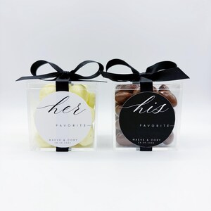 May include: Two clear square boxes filled with candies, each tied with a black ribbon. The left box has a white circular label reading her FAVORITE MAEVE & CORY 28.09.2026. The right box has a black circular label reading his FAVORITE MAEVE & CORY 28.09.2026.