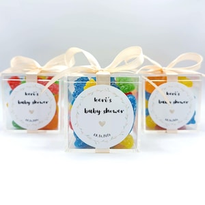 May include: Three clear square plastic favor boxes filled with colorful gummy candies. Each box is tied with a cream-colored ribbon and features a circular white label printed with the text Keri's baby shower, a small heart, and the date 03.16.2026.