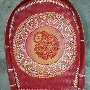 May include: A hand-woven winnowing tray made of bamboo, painted in a vibrant red with a central circular design. The center features a yellow-to-orange gradient background with a detailed red paisley motif, surrounded by white leaf patterns.