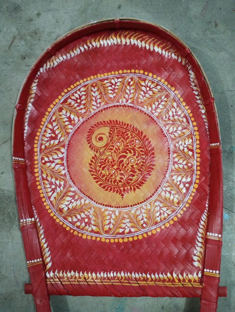 Puede incluir: Una bandeja aventadora tejida a mano en bamb&uacute;, pintada en rojo vibrante con un dise&ntilde;o circular central. El centro presenta un fondo degradado de amarillo a naranja con un motivo de cachemira rojo detallado, rodeado de patrones de hojas blancas.