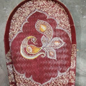 May include: A hand-woven winnowing tray made of bamboo, featuring a deep red stained finish. The surface is decorated with intricate cream-colored floral patterns and a central stylized peacock motif in shades of yellow, orange, and white.