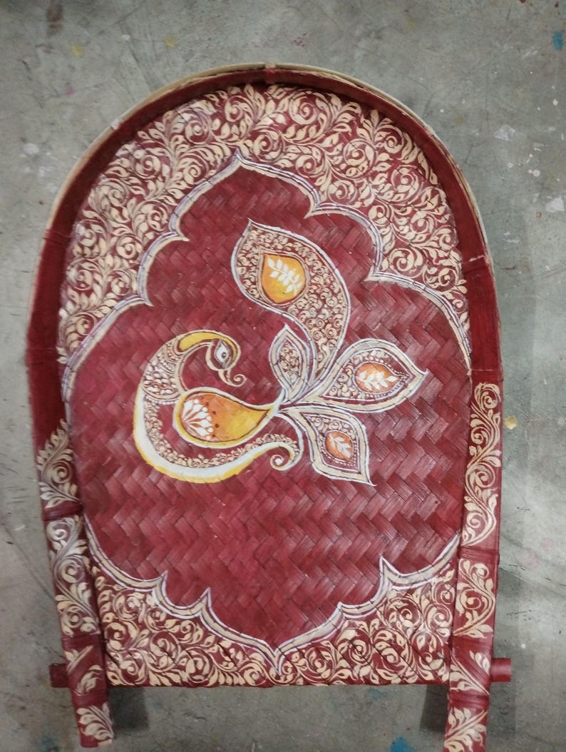 May include: A hand-woven winnowing tray made of bamboo, featuring a deep red stained finish. The surface is decorated with intricate cream-colored floral patterns and a central stylized peacock motif in shades of yellow, orange, and white.