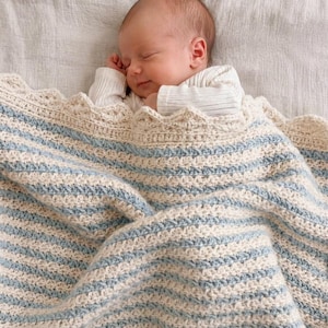 May include: A newborn infant with light skin and short hair is sleeping peacefully while wrapped in a handmade crochet blanket. The blanket features alternating horizontal stripes of cream and light blue yarn with a scalloped decorative edge.