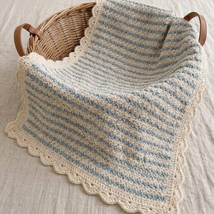 May include: A crochet blanket featuring alternating horizontal stripes of cream and light blue yarn. The blanket has a textured stitch pattern and is finished with a decorative scalloped cream border. It is draped over a round woven wicker basket.
