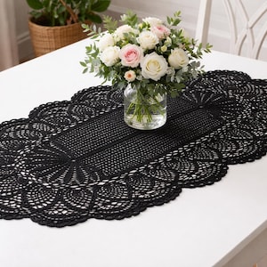 Peut inclure: Une nappe ovale en dentelle noire avec un bord festonné est posée sur une table blanche. Un vase en verre avec des roses blanches et roses et de la verdure est centré sur la nappe. La table est près d'une fenêtre.