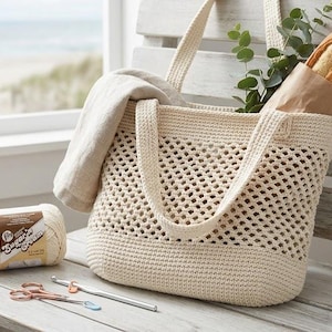 May include: A cream-colored, crocheted tote bag with a rectangular base and open weave design. The bag contains a folded linen cloth, a baguette in a paper sleeve, and greenery. A ball of cream-colored yarn, scissors, and crochet hooks are on the bench.