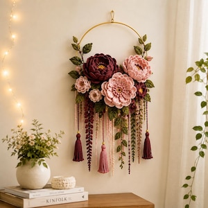 Puede incluir: Un adorno de pared que consiste en un aro de metal dorado decorado con flores de ganchillo en tonos burdeos intenso, rosa suave y rosa claro. Hojas verdes de ganchillo y largos hilos colgantes con borlas a juego cuelgan del aro.