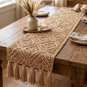 May include: A beige crochet table runner featuring a repeating diamond pattern and finished with long fringe tassels at the ends. The runner is laid out on a rustic wooden dining table, accompanied by a ceramic vase with dried grass and a candle.