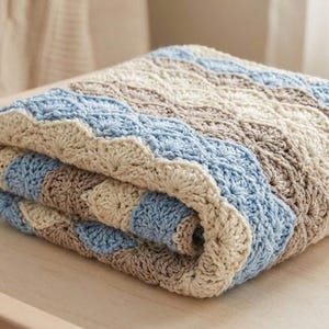 Könnte beinhalten: Eine gefaltete, gehäkelte Babydecke in Blau-, Beige- und Brauntönen. Die Decke hat einen gewellten Rand und ein strukturiertes, wellenartiges Muster. Ein Baby-Beißring aus Holz und ein kleines Stofftier liegen daneben.