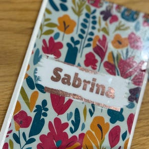 May include: A notebook with the name "Sabrina" in rose gold lettering. The cover displays a floral pattern with red, orange, teal, and purple flowers on a light green backdrop. The notebook has a white spine.