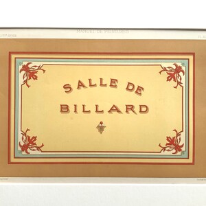 以下が含まれることがあります： クリーム色の背景に赤色の「SALLE DE BILLARD」の文字が中央に配置された、額装されたヴィンテージプリント。プリントには茶色の縁取りがあり、角には赤と緑の花柄のアクセントがあります。上部には「MANUEL DE PEINTURES」と書かれています。