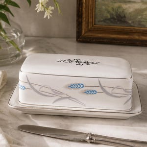 May include: A white ceramic butter dish with a rectangular lid and base, featuring a silver trim, a black decorative flourish on top, and a pattern of blue and grey wheat stalks along the sides, accompanied by a silver-toned metal butter knife.