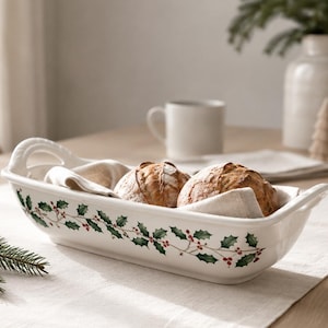 May include: A rectangular white ceramic serving dish with two handles, featuring a painted green holly leaf and red berry pattern along the exterior sides. The dish contains two round crusty bread loaves nestled inside a folded beige linen cloth.