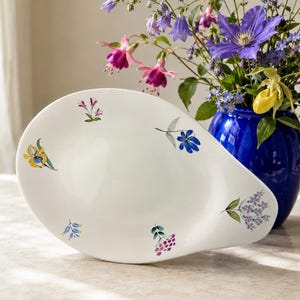 May include: A white, oval-shaped ceramic serving platter featuring a handle on one side and delicate, hand-painted botanical illustrations of various small flowers and berries scattered across the surface in shades of blue, yellow, pink, and green.