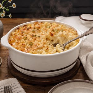May include: A round white ceramic casserole dish with a dark brown horizontal stripe, filled with baked macaroni and cheese topped with herbs and black pepper, featuring a matching lid and a silver metal serving spoon resting inside the dish.