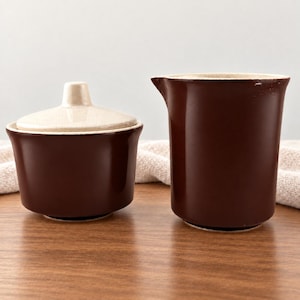 May include: A dark brown ceramic sugar bowl with a matching cream-colored lid and a separate dark brown ceramic creamer pitcher. Both pieces feature a smooth, glossy finish and a simple, functional design for serving coffee or tea condiments.