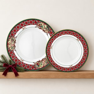 May include: Two round white ceramic plates featuring a red and green plaid border with gold trim. The rims are decorated with illustrations of small animals, birds, and fruit clusters. A dark red velvet bow sits near the plates.