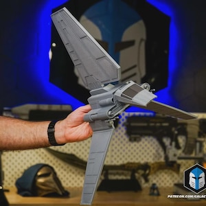 May include: A gray plastic model of a Lambda-class shuttle spacecraft featuring a tall central vertical stabilizer wing, two smaller side wings, a cockpit with dark windows, and cylindrical engine components on the main fuselage body.