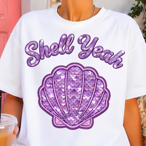 Puede incluir: Camiseta blanca con el texto "Shell Yeah" en fuente morada brillante sobre un diseño de concha morada con lentejuelas. Una prenda casual para el día a día.