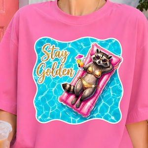May include: Pink t-shirt featuring a cartoon raccoon wearing sunglasses and a bikini, lounging on a pink pool float in a pool. The text "Stay Golden" is written in gold script above the raccoon.