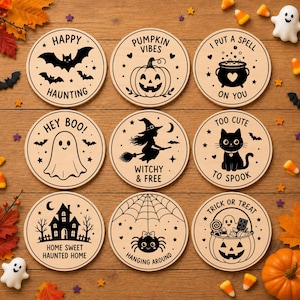 May include: Nine circular wooden coasters featuring black Halloween-themed illustrations and text. Designs include a bat with 'HAPPY HAUNTING', a pumpkin with 'PUMPKIN VIBES', a cauldron with 'I PUT A SPELL ON YOU', a ghost with 'HEY BOO!', a witch with 'WITCHY & FREE', a cat with 'TOO CUTE TO SPOOK', a house with 'HOME SWEET HAUNTED HOME', a spider with 'HANGING AROUND', and a candy bucket with 'TRICK OR TREAT'.