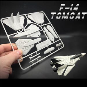 Può includere: Un kit di modellismo F-14 Tomcat bianco con dettagli neri, parzialmente assemblato. Il kit include varie parti non assemblate su un telaio di plastica, e il modello finito è esposto accanto. Il testo "F-14 TOMCAT" è visibile.