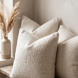 May include: Four cream-colored, textured throw pillows arranged on a rustic wooden bench. The pillows have a soft, plush appearance. A vase with dried plants sits nearby, adding a touch of natural decor. The pillows are a neutral color.