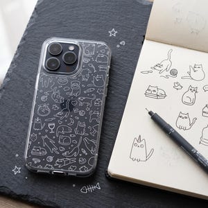 Puede incluir: Una funda transparente para smartphone con un patrón repetitivo de líneas blancas que muestra varios gatos, espinas de pescado, ovillos de lana y pequeñas estrellas, sobre una superficie oscura texturizada junto a un cuaderno de bocetos.