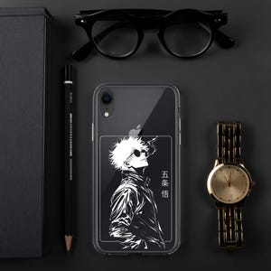 Puede incluir: Una funda transparente para teléfono con una ilustración de anime en blanco y negro de un personaje con gafas de sol y el texto "五条 悟". La funda está junto a un reloj dorado, gafas negras, un lápiz y un cuaderno negro.