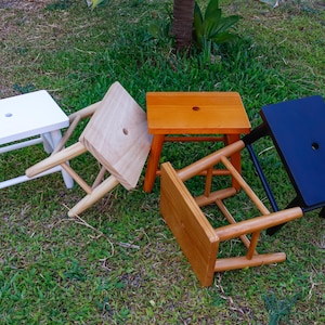 Peut inclure: Quatre petits tabourets en bois avec des plateaux rectangulaires, chacun doté d'un trou circulaire central. Les tabourets sont finis en blanc, bois clair naturel, brun moyen et noir, chacun soutenu par quatre pieds avec traverses.