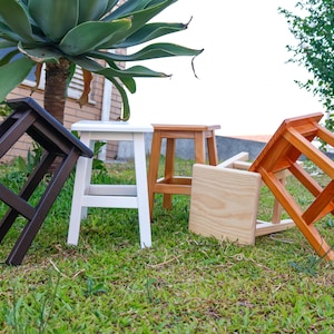 May include: A collection of four wooden stools in various finishes, including dark brown, white, natural light wood, and a warm orange-toned wood. The stools feature four legs and a flat square seat, arranged in a group on a grassy lawn area.