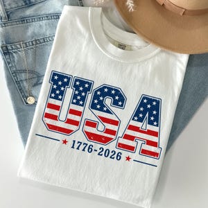 A white t-shirt with the text "USA" printed on it, along with the year "1776-2026" below it. The t-shirt is laid out on a surface, with a beige hat and some jeans visible in the background.