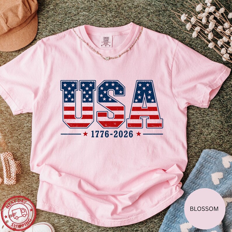 a pink t-shirt with the text "USA" and "1776-2026" printed on it, along with an American flag design. The t-shirt is laid out on a surface, surrounded by various items such as a hat, a necklace, and a blue towel.