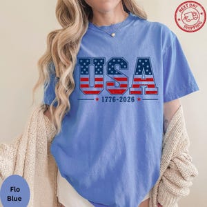 A woman wearing a blue t-shirt with the text "USA" printed on it, along with the date "1776-2026" below it. She is also wearing a beige cardigan.