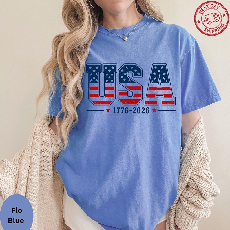 A woman wearing a blue t-shirt with the text "USA" printed on it, along with the date "1776-2026" below it. She is also wearing a beige cardigan.