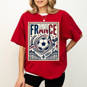A woman wearing a red t-shirt with a graphic design featuring a soccer ball, buildings, and a rooster, along with the word "FRANCE" prominently displayed.